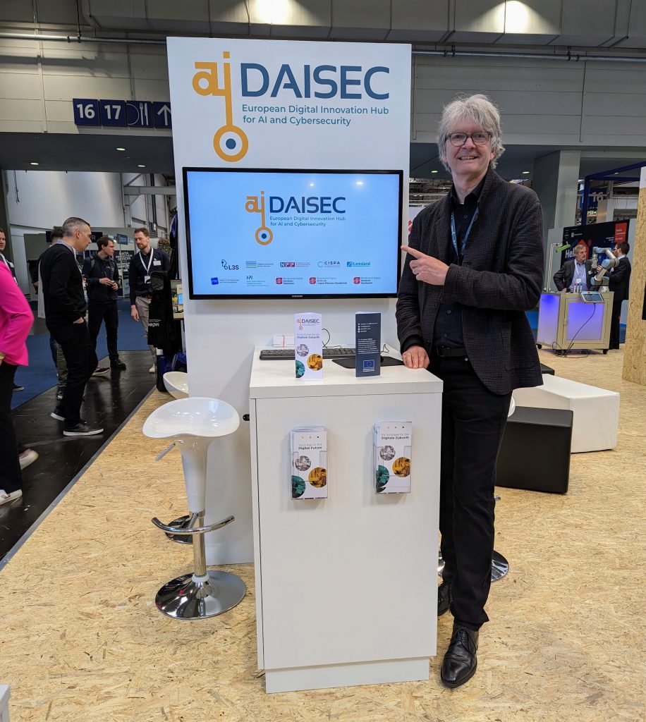 Walter Pirk steht rechts neben dem DAISEC Stand mit dem DAISEC Logo und präsentiert den DAISEC Film auf einem Bildschirm am Stand