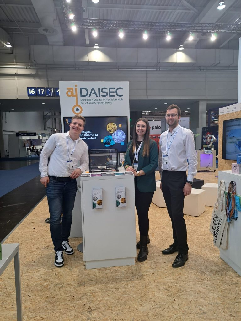 Timo Bartels, Nicole Bartodziej und Karl Hilbig am DAISEC Stand mit Flyern und einer DAISEC Präsentation auf einem Bildschirm